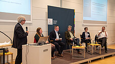 Podiumsdiskussion mit sechs Personen auf der Bühne, Moderator am Rednerpult