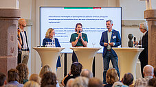 Podiumsdiskussion mit fünf Personen vor einer Leinwand, im Vordergrund sitzendes Publikum