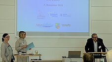 Zwei Personen stehen vor einer Präsentation mit dem Titel Nachhaltige Kommunen in Brandenburg: Wie weiter?, eine dritte Person spricht am Rednerpult