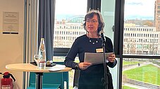 Person spricht an einem Mikrofonstativ und hält Papiere; im Hintergrund großes Fenster mit Blick auf eine Stadtansicht