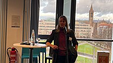 Person spricht an einem Mikrofonstativ neben einem Stehtisch; hinter ihr Stadtpanorama mit Kirchturm durch ein großes Fenster