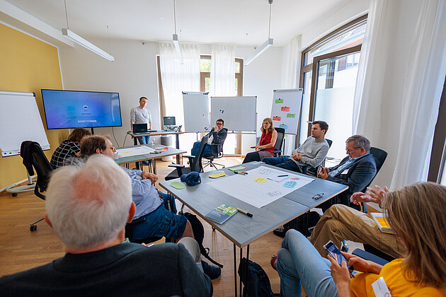 Gruppe sitzt im Halbkreis in einem Workshop-Raum, eine Person präsentiert vor Flipchart und Bildschirm