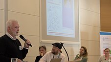 Person spricht mit Mikrofon vor Publikum und Präsentationsfolie mit Karte im Hintergrund