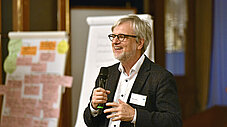 Person spricht lachend in ein Mikrofon bei einer Veranstaltung, im Hintergrund Flipchart-Tafeln mit Notizen
