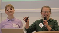Zwei Personen an einem Podium: eine spricht ins Mikrofon, die andere hält eine Fernbedienung