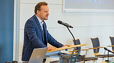 Mann im karierten blauen Sakko spricht an Rednerpult mit Mikrofon vor einer Leinwand