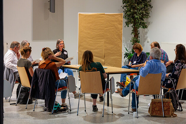 Gruppe von etwa zehn Personen diskutiert an einem Tisch, eine Person präsentiert mit Unterlagen in der Hand