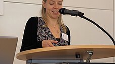 Person spricht lächelnd an einem Redepult mit Mikrofon, Pult trägt Aufschrift Heimvolkshochschule am Seddiner See