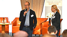Zwei Personen stehen vor einem Publikum, eine spricht ins Mikrofon, im Hintergrund orangefarbene Sessel auf einer Bühne