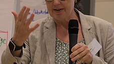 Person spricht mit Mikrofon in der Hand und erhobener Hand bei einer Veranstaltung, Flipchart im Hintergrund