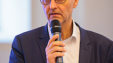 Person spricht ins Mikrofon bei einer Veranstaltung, Namensschild am Revers sichtbar