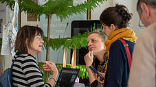 Drei Personen im Gespräch an einem Ausstellungsstand mit Laptop und Pflanzen