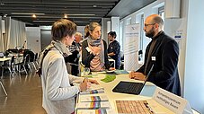 Zwei Besucherinnen im Gespräch mit einem Aussteller am Stand des Kompetenzzentrums nachhaltiges Bauen mit Broschüren und Laptop