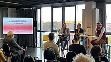 Drei Personen auf einem Podium diskutieren; eine Leinwand zeigt "Nachhaltige Kommunen in Brandenburg 2023: Wie weiter?"; Publikum im Vordergrund