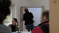 Person schreibt auf ein Flipchart, Teilnehmende sitzen an einem Tisch im Workshopraum