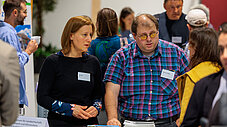 Frau und Mann mit Namensschildern schauen gemeinsam auf Materialien an einem Messestand, im Hintergrund weitere Veranstaltungsgäste.