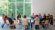 Gruppe von Personen sitzt in einem Stuhlkreis zur Diskussion in einem hellen Raum mit Fensterfronten und Baumblick.