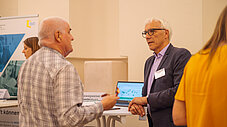 Zwei Personen im Gespräch an einem Ausstellungsstand mit Laptop und Roll-up
