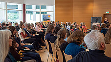 Volles Publikum sitzt in Reihen bei einer Veranstaltung, Rednerin am rechten Rand