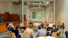 Etwa 15 Personen sitzen in einem Stuhlkreis in einem hellen Saal mit Industriedecke und diskutieren miteinander.