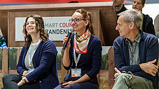 Drei Personen sitzen bei einer Podiumsdiskussion vor Smart Country Brandenburg Kulisse; eine Person spricht ins Mikrofon