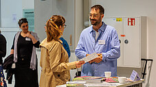 Zwei Personen tauschen an einem Infotisch Flyer aus, im Hintergrund weitere Veranstaltungsgäste