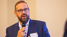 Person spricht ins Mikrofon bei einer Veranstaltung, Namensschild am Revers