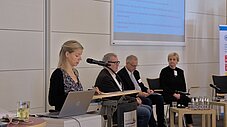 Person spricht am Rednerpult mit Laptop und Mikrofon, drei Personen sitzen auf dem Podium, Präsentationsfolie im Hintergrund