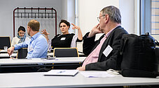 Gruppe diskutiert an Tischen in einem Seminarraum, Person gestikuliert