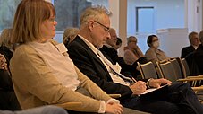 Zwei Personen im Vordergrund sitzen bei einer Konferenz, eine macht Notizen, weitere Zuhörende im Hintergrund