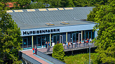 Gebäude der Hufeisenfabrik mit Metallschriftzug, umgeben von Bäumen, Menschen auf der Außenterrasse bei Sonnenschein.