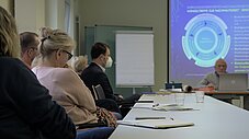 Teilnehmende sitzen an einem langen Tisch im Seminarraum, Person präsentiert vorne, Kreisdiagramm auf Leinwand