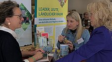 Drei Personen im Gespräch an einem Messestand mit Banner "LEADER, Leute, Ländlicher Raum" und Informationsmaterial