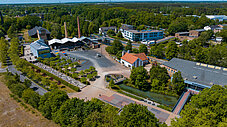 Luftaufnahme eines Geländes mit Industriegebäuden, Schornstein, gepflasterten Wegen und umgebender Baumlandschaft.