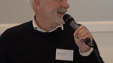 Person spricht lächelnd ins Mikrofon vor einer Präsentationsleinwand
