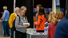 Lächelnde Frau mit grauem Haar und Namensschild steht an einem Ausstellungsstand und hält Informationsmaterialien in der Hand.