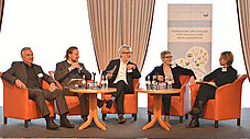 Fünf Personen sitzen auf einer Bühne in orangefarbenen Sesseln bei einer Podiumsdiskussion