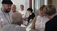 Gruppe von Personen im Gespräch an einem Tisch mit Namensschildern, darunter agrathaer