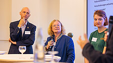 Drei Personen bei Podiumsdiskussion, eine spricht ins Mikrofon, Projektionsscreen im Hintergrund