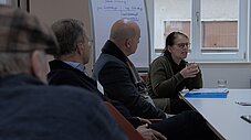 Kleingruppe sitzt an einem Tisch in einem Workshop, eine Person spricht mit Gesten, im Hintergrund ein beschriebenes Flipchart