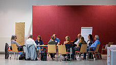 Rund zehn Personen sitzen in einem Halbkreis an Tischen in einem hellen Saal mit roter Wand und diskutieren; ein Flipchart steht im Hintergrund. (Rechte: @MLEUV)