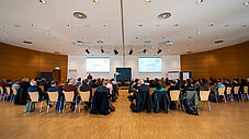 Großer Konferenzsaal mit Publikum in Halbkreisanordnung vor zwei Leinwänden, Redner vorne