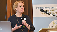Person spricht in ein Mikrofon am Rednerpult, im Hintergrund Banner mit Aufschrift zur Nachhaltigkeitsforschung