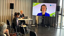 Publikum verfolgt eine Videokonferenz auf einer großen Leinwand mit ZWK-Logo; Moderatorin sitzt an einem Tisch mit Laptop