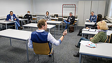 Gruppe diskutiert im Stuhlkreis in einem Seminarraum, Moderator gestikuliert
