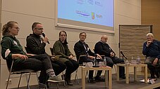 Sechs Personen sitzen auf einer Bühne als Podium, eine spricht ins Mikrofon, im Hintergrund eine Projektionsleinwand