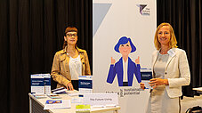 Zwei Frauen stehen am Stand von The Future Living vor einem Rollup-Banner mit dem Slogan Unbox your sustainable potential und Informationsmaterial.