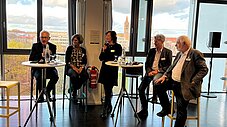 Fünf Personen beim Podiumsgespräch auf Barhockern vor großen Fenstern mit Stadtblick, eine Person spricht ins Mikrofon