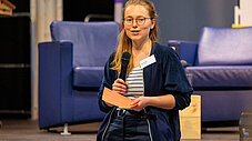 Junge Frau mit Brille und Namensschild spricht mit Mikrofon in der Hand vor blauen Sofas bei einer Veranstaltung.