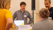 Gruppe steht im Gespräch an einem Informationsstand mit Laptop und Ausstellungsmaterialien